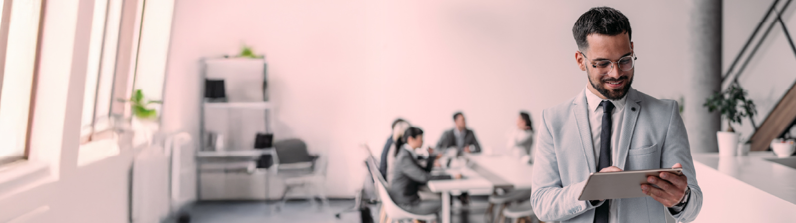 A professional in business attire stands in focus with a tablet, while a group of colleagues collaborate at a conference table in the blurred background.