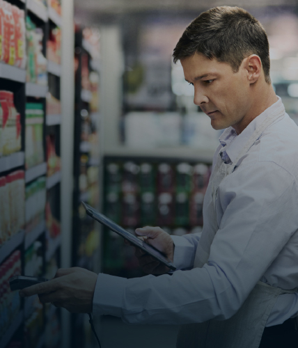 A retail store employee scans products on a supermarket shelf using a handheld device while holding a tablet, surrounded by neatly arranged grocery items.
