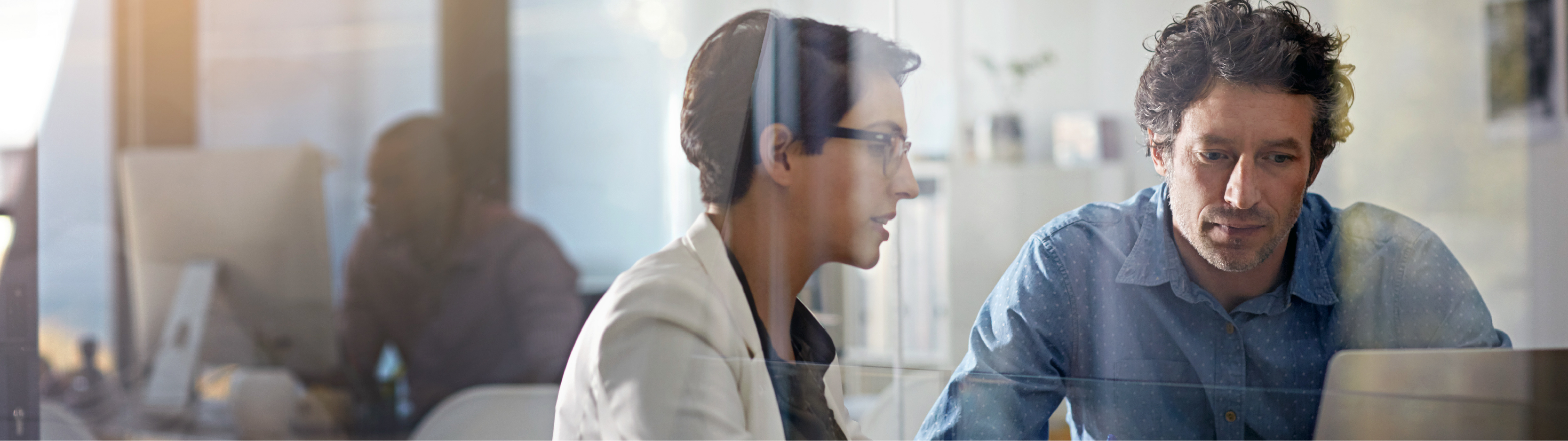 Two professionals reviewing data together on a laptop in a modern office environment, viewed through a glass wall.