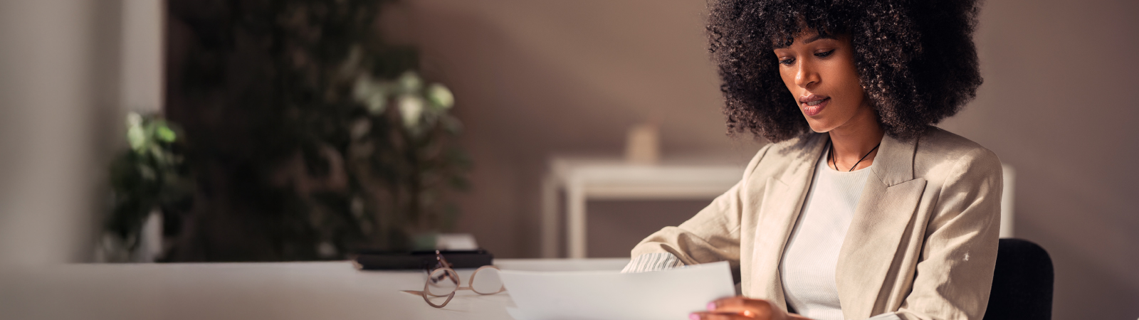 A woman in a beige blazer sits at a desk reviewing documents in a softly lit office, with reading glasses on the table and a blurred plant in the background.
