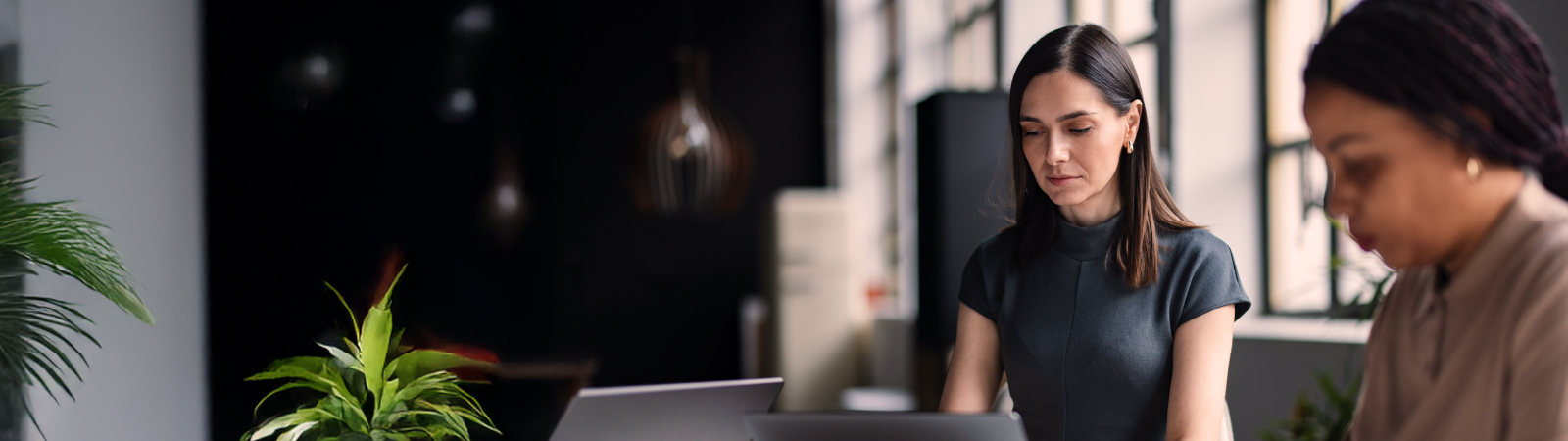 A focused woman in a dark dress works on a laptop at a modern office table, with another colleague blurred in the foreground and natural light streaming through large windows.