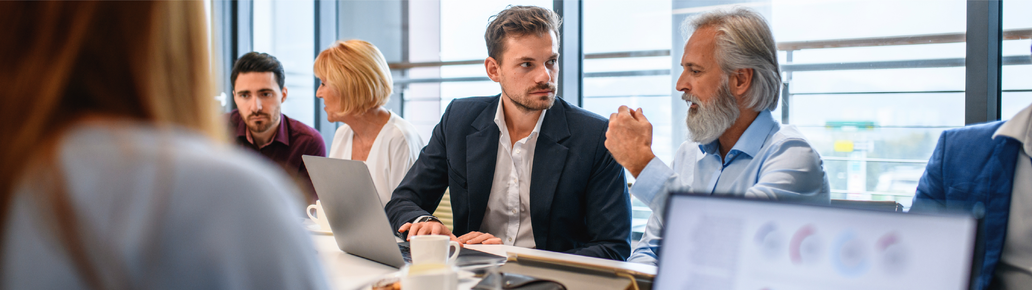 Business professionals in a meeting room engaged in discussion, with charts displayed on a laptop in the foreground.