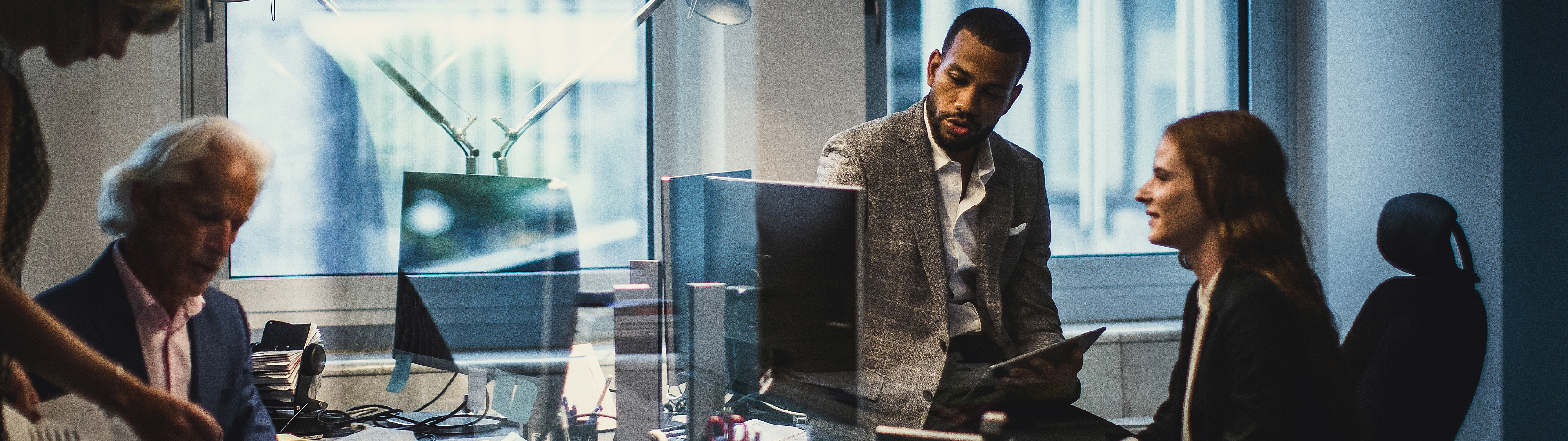 Three professionals in a modern office collaborating around computer monitors and documents, with reflections on glass walls creating a dynamic workspace atmosphere.