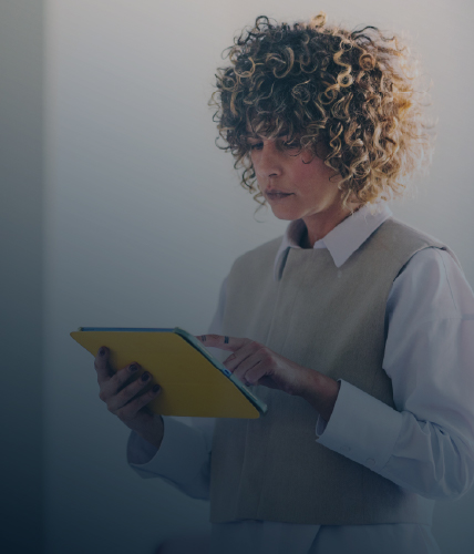 A close-up of a woman with curly hair concentrating on a yellow tablet in a softly lit office.