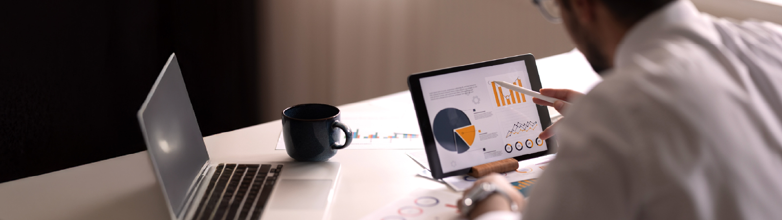 A man using a digital tablet to review financial pie charts, bar graphs, and trend lines at a desk with printed reports, a laptop, and a coffee mug.