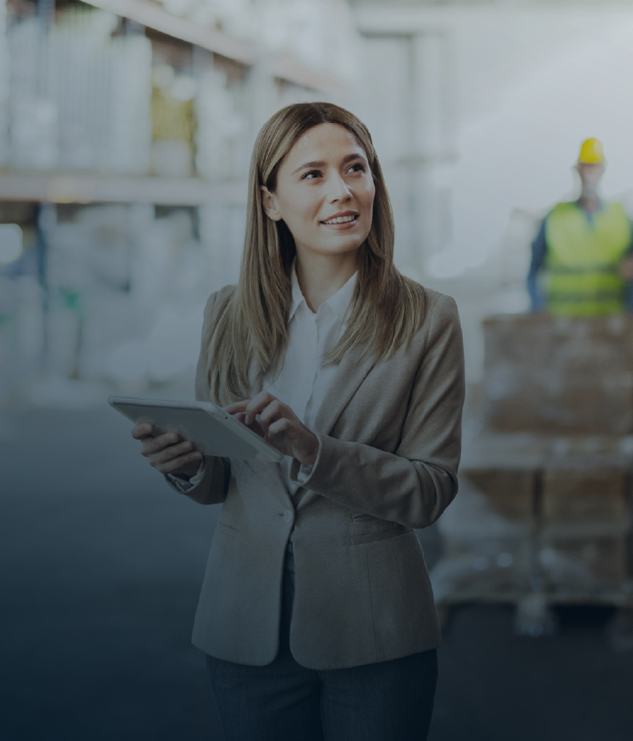 A business professional using a tablet inside a warehouse, with inventory shelves and workers wearing safety vests visible in the background.