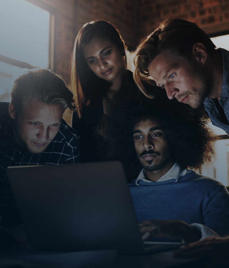 Four professionals gathered closely around a laptop in a modern office with exposed brick walls and warm lighting, focused on the screen as they collaborate.