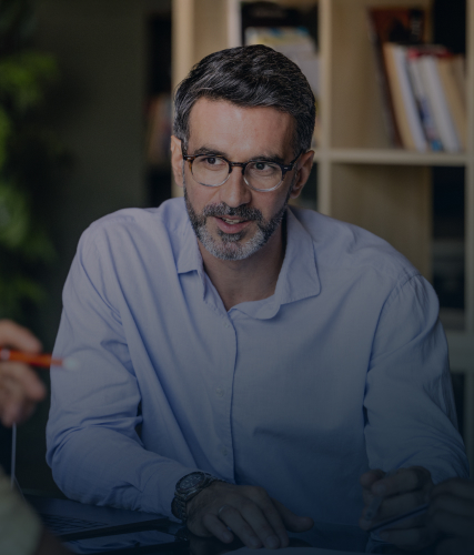 A business professional wearing glasses speaking during a meeting while holding a pen, seated at a table with colleagues in an office workspace.