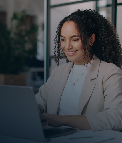 A professional woman smiling while working on a laptop at a desk in a modern office environment.