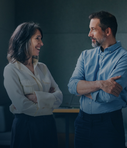 Two colleagues standing face-to-face in conversation, smiling, with arms crossed in a casual office setting.