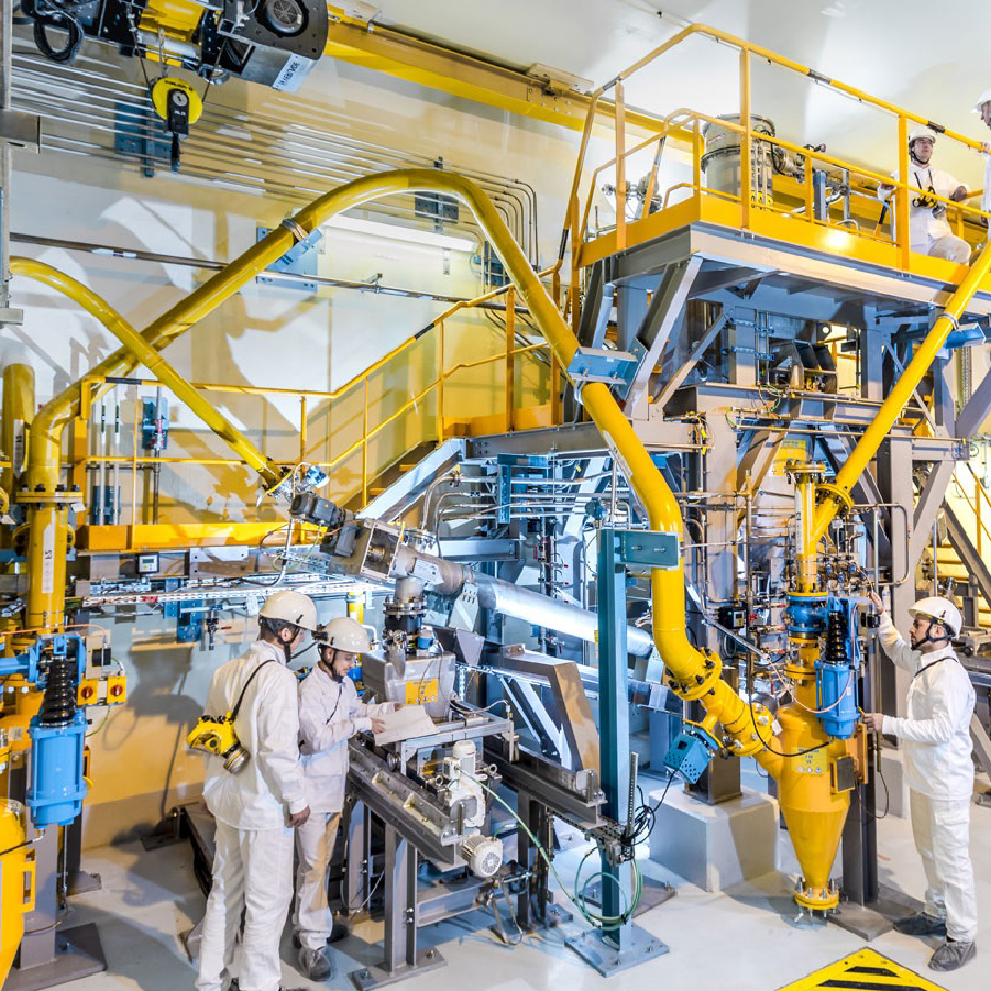 Workers in protective clothing operating machinery inside a brightly lit industrial facility with large yellow pipes and equipment.