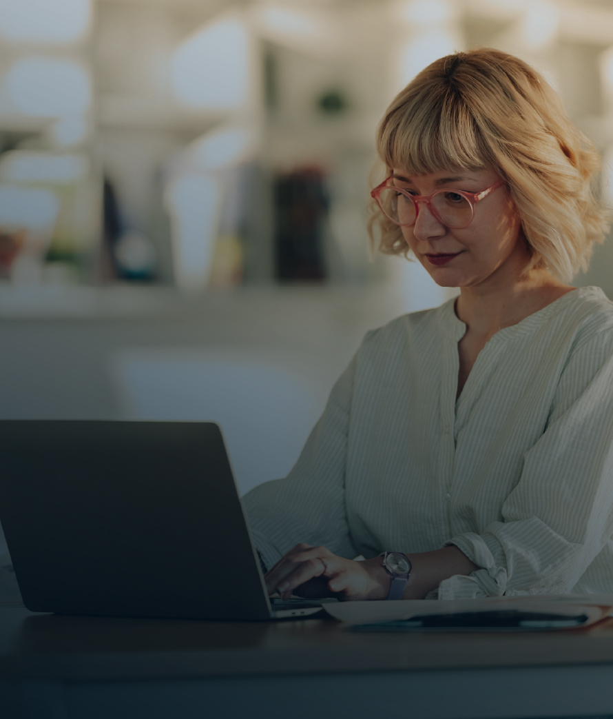 A professional woman wearing glasses reviews notes while working on a laptop in a contemporary office setting with soft lighting and blurred background details.