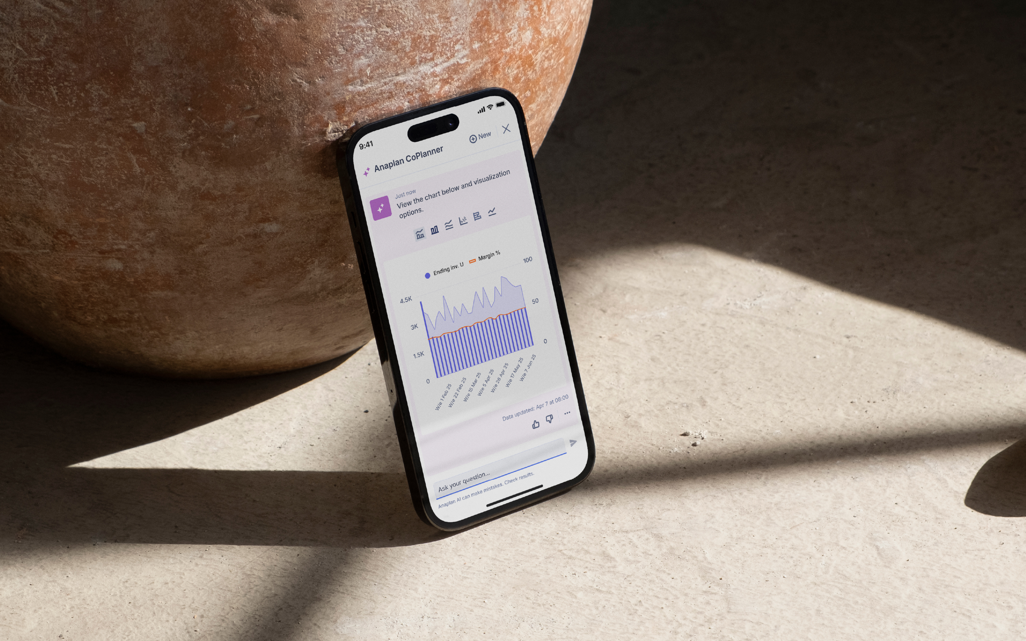 A smartphone leaning against a large clay pot displays the Anaplan CoPlanner mobile interface with a chart showing Ending Inv. and Margin % trends, placed on a sunlit concrete surface with shadows cast across the floor.