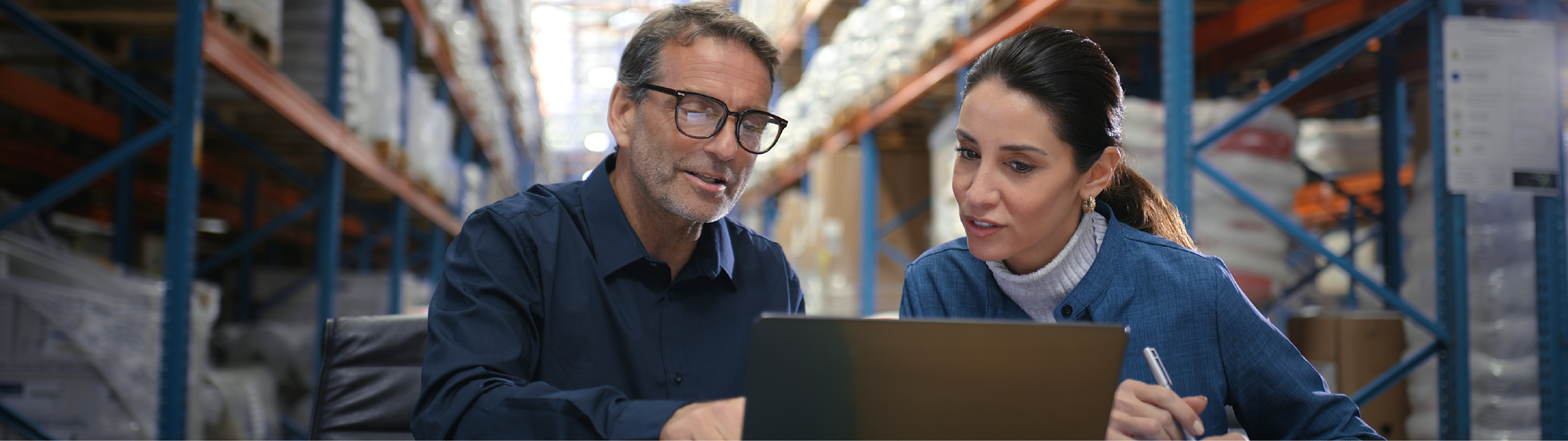 Man and woman seated at a desk in a warehouse, reviewing information on a laptop together.