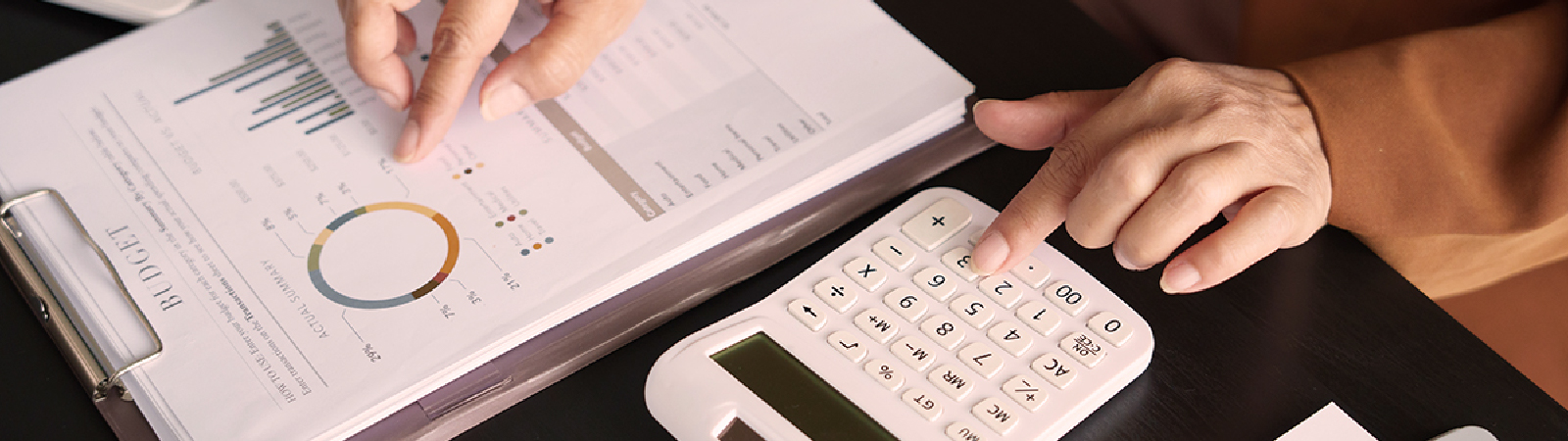 A close-up of a person's hands working on financial documents, pointing at a budget report with charts and graphs while using a white calculator on a dark desk.