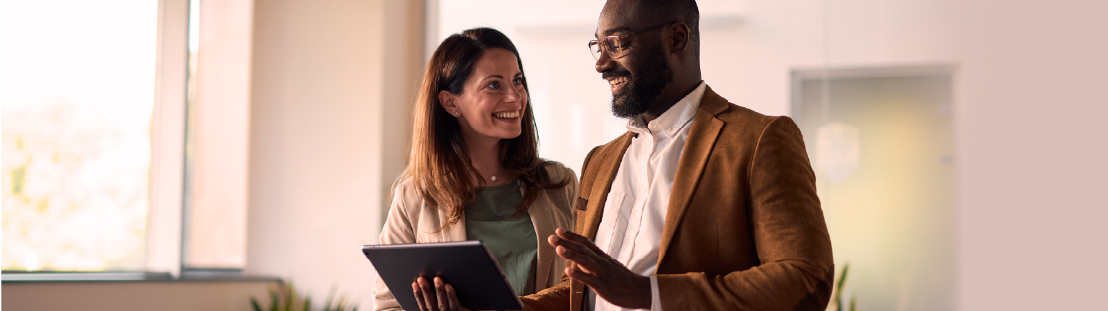 Two colleagues standing in a conference room, smiling and conversing while one holds a tablet.