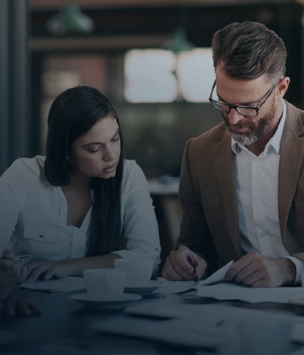Two professionals seated at a table reviewing documents together in a modern office setting. One person writes notes while the other observes attentively.