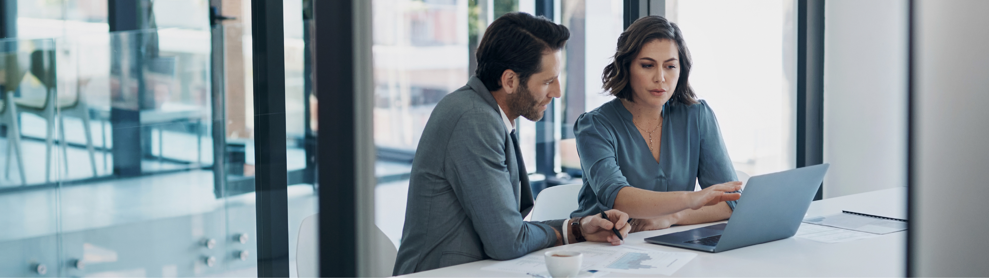 Two coworkers sitting at a table in an office talking over a laptop together