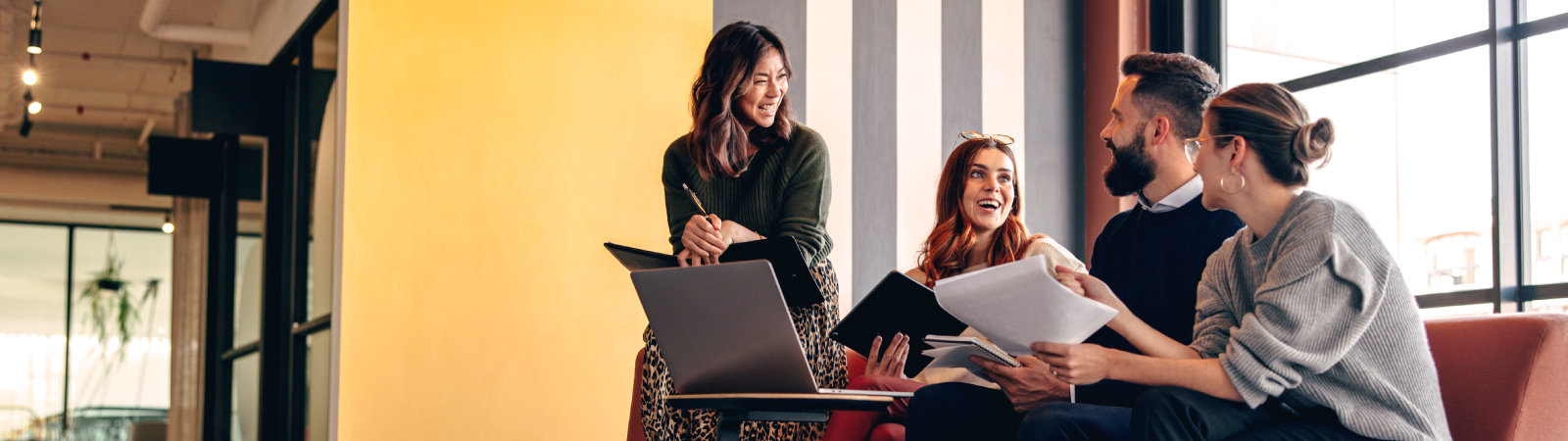 Group of four young professionals sitting in a modern office lounge, smiling and collaborating with notebooks and a laptop in front of a striped wall and large windows.