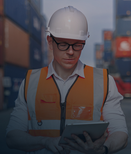 A worker wearing a hard hat and orange safety vest standing in a shipping yard, looking at a tablet with stacked cargo containers behind him.