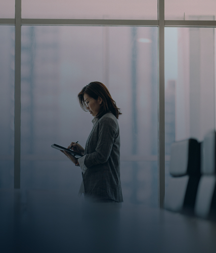 A professional woman standing in a glass-walled office, reviewing notes on a tablet, with office chairs and a city skyline visible in the background.