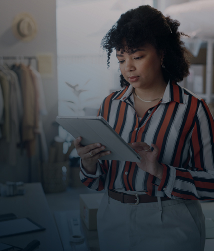 A woman in a striped shirt holding a tablet and reviewing information in a retail workspace with clothing racks and packaging materials in the background.
