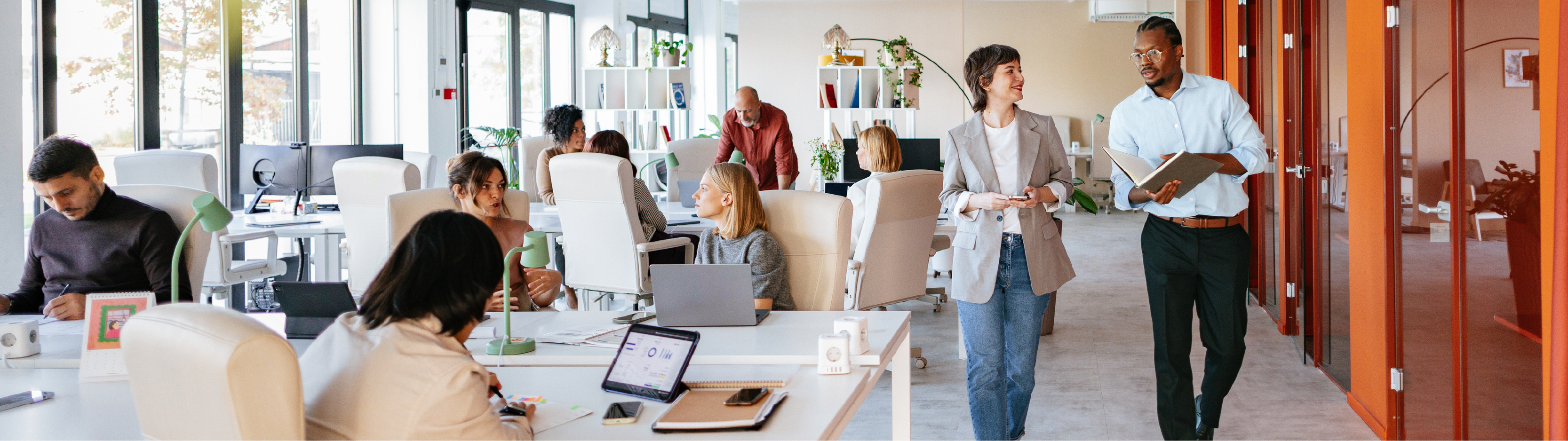 Modern open-plan office with professionals working at desks and in discussion. Two colleagues walk together in conversation near a hallway with glass and orange-framed walls.