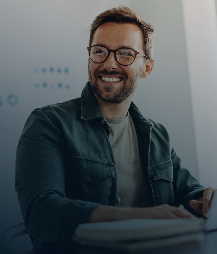A professional smiling while working on a laptop at a desk in a modern office environment.