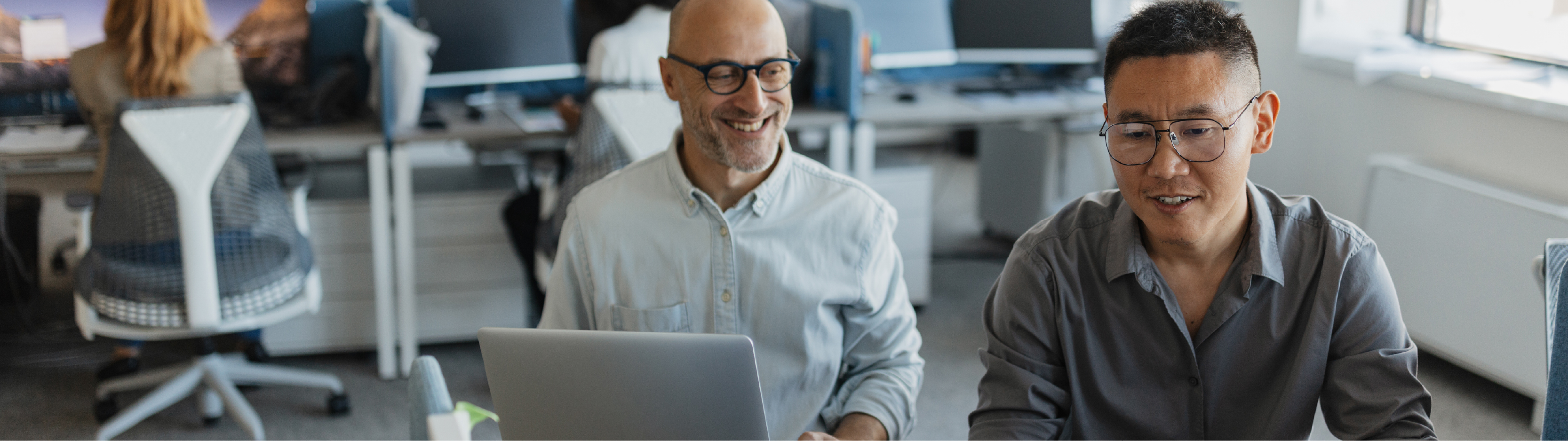 Two male coworkers smiling and working together in a collaborative office space, with laptops and desktop monitors on a shared desk.