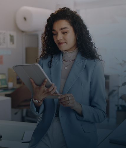 Woman in a light blue blazer stands in an office workspace, looking at and tapping a tablet.
