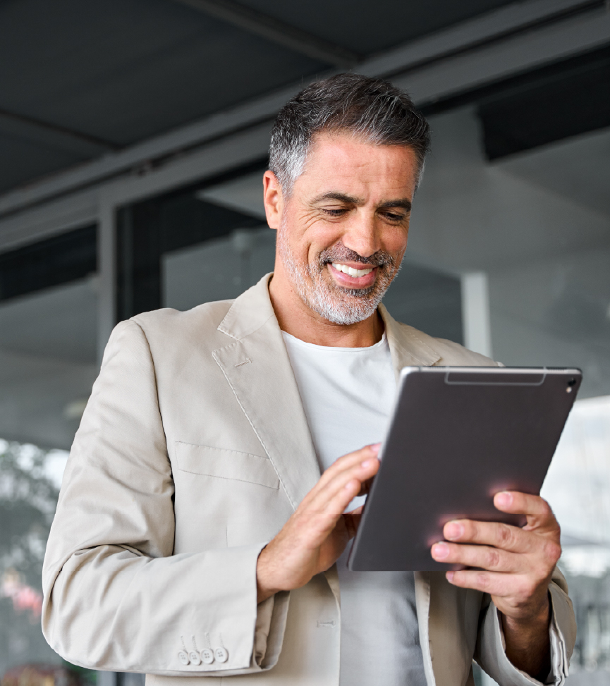 A person in a beige blazer smiles while holding and tapping on a tablet outdoors near a glass building.