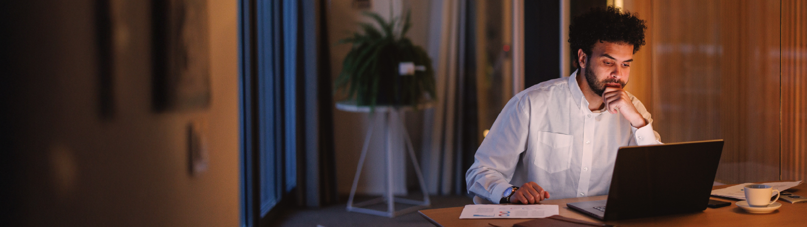 A man in a white shirt working at a desk in a dimly lit office, focused on a laptop screen. Papers, a notebook, and a cup of coffee are visible on the table.