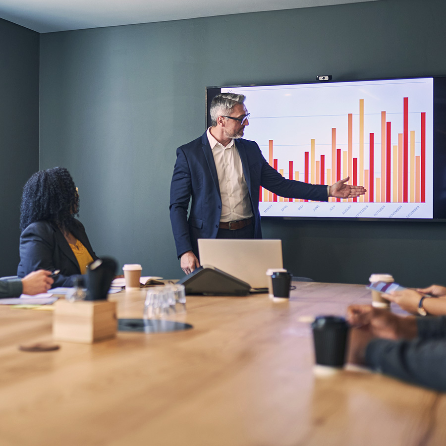 A business meeting where a presenter points to a bar chart on a wall-mounted screen while colleagues sit around a conference table with laptops and coffee cups.