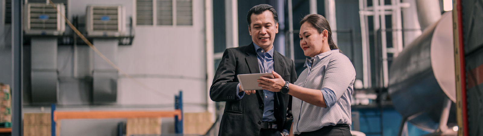 Two professionals standing in an industrial warehouse reviewing a tablet together.