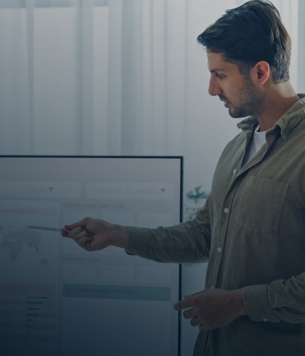 Man standing and pointing at a presentation screen displaying charts and analytics data in a softly lit office setting.