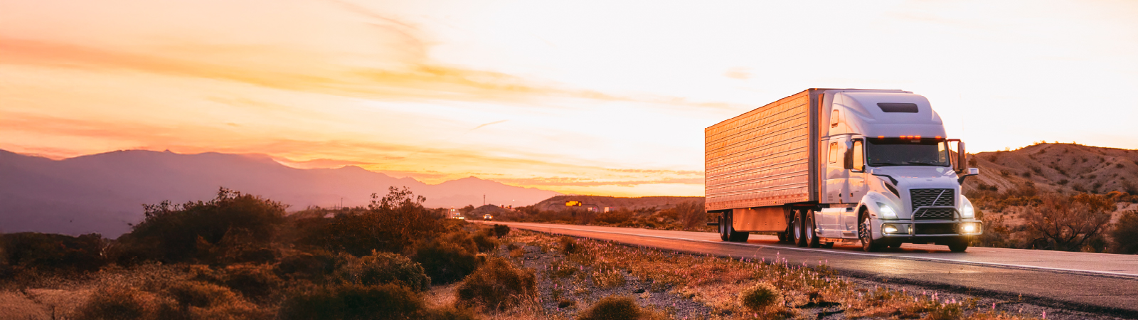 A white semi-truck drives along a desert highway at sunset, with golden light reflecting off its trailer and surrounding landscape.