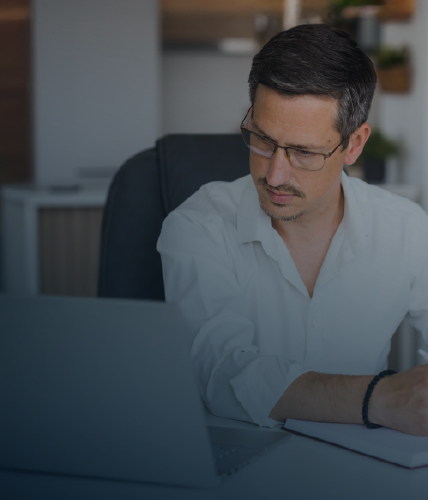 A professional in a white shirt works at a desk, taking notes while looking at a laptop in a contemporary office setting.