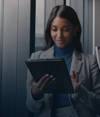 A woman in a business suit standing near large windows, holding and using a tablet with a focused expression.