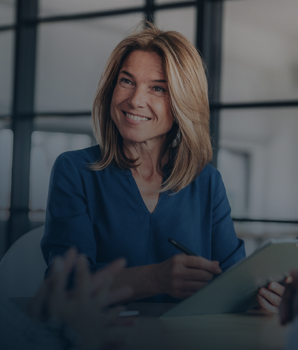 A business professional smiling during a meeting, holding a tablet while seated at a conference table in a modern office setting.
