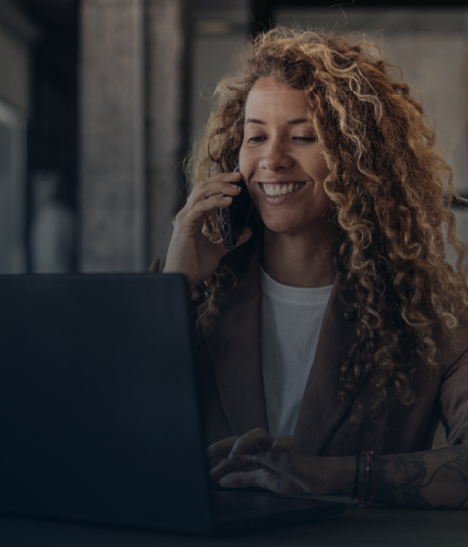 A woman with curly hair smiling while talking on the phone and working on a laptop at a wooden desk in an office setting.