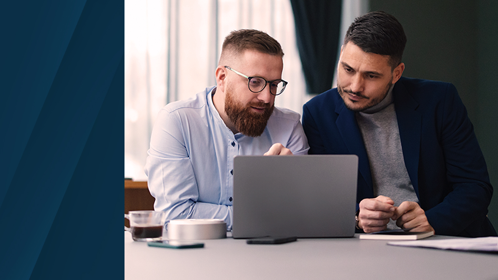 Two professionals sitting at a table, reviewing data on a laptop and discussing work in a well-lit office environment.