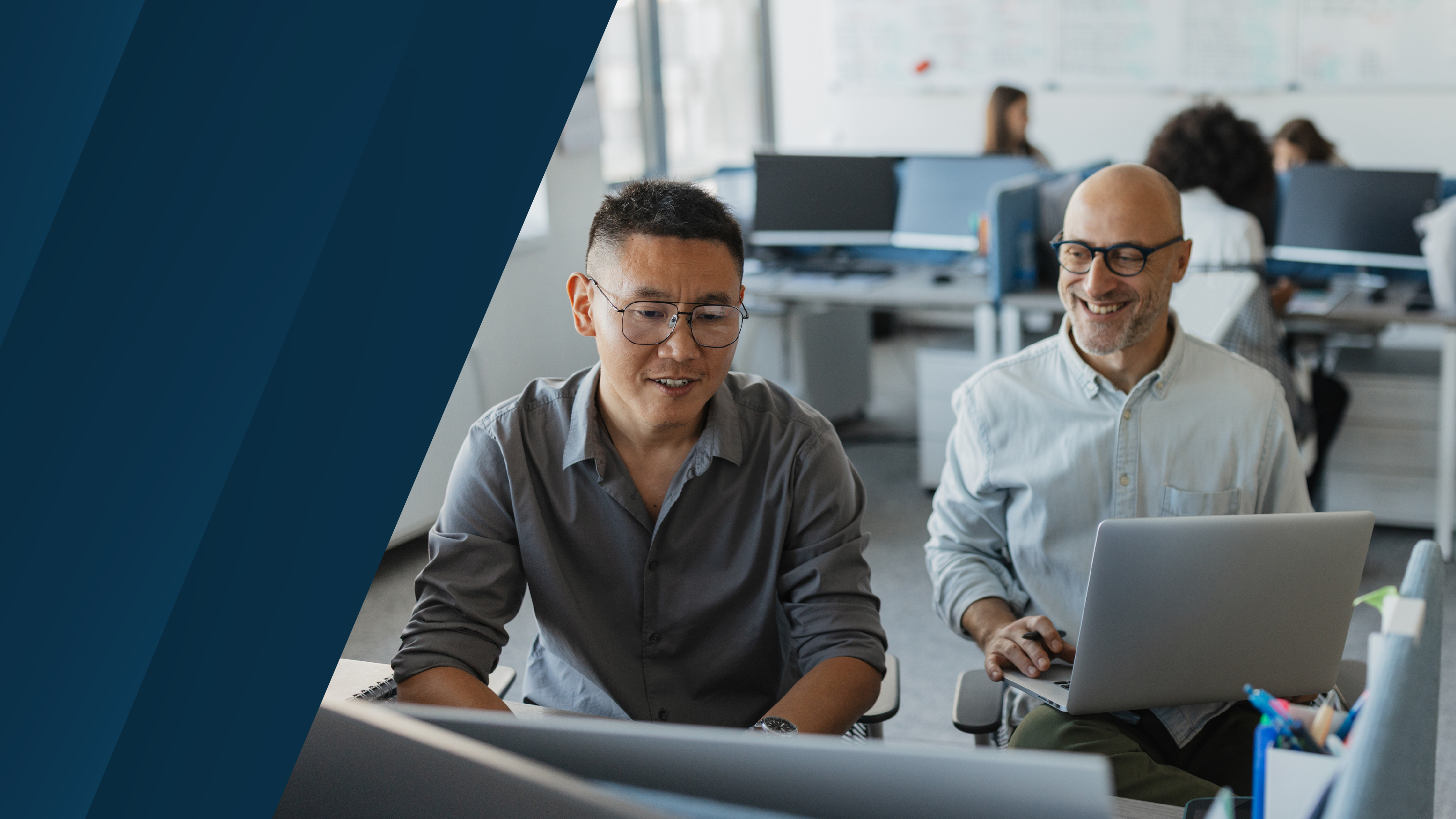 Two professionals working together in a modern open office environment, one using a desktop monitor while the other smiles and works on a laptop, with dark blue diagonal graphic panels on the left.