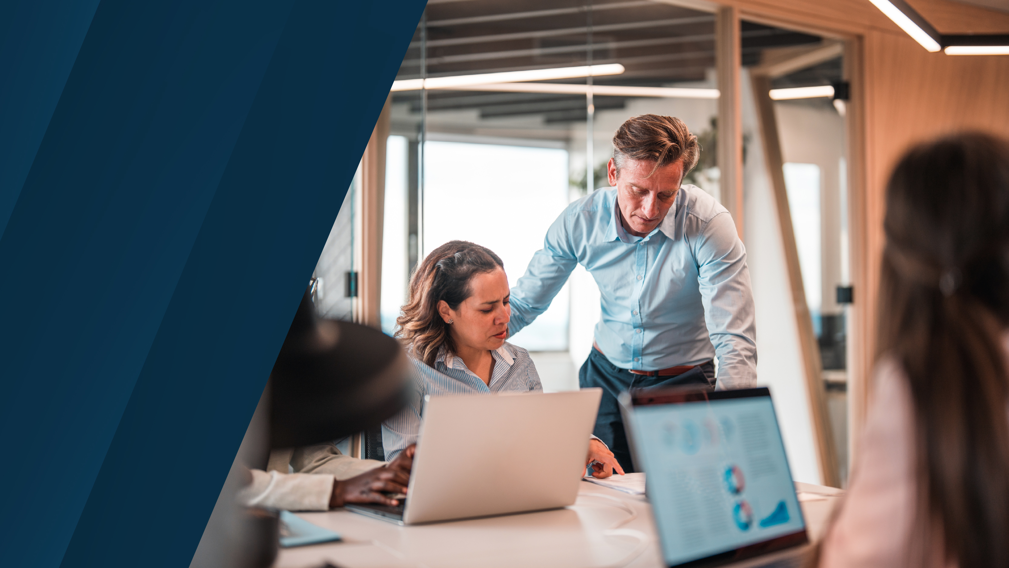Business professionals working together around a conference table with laptops and documents, engaged in discussion within a modern, glass-walled meeting room.