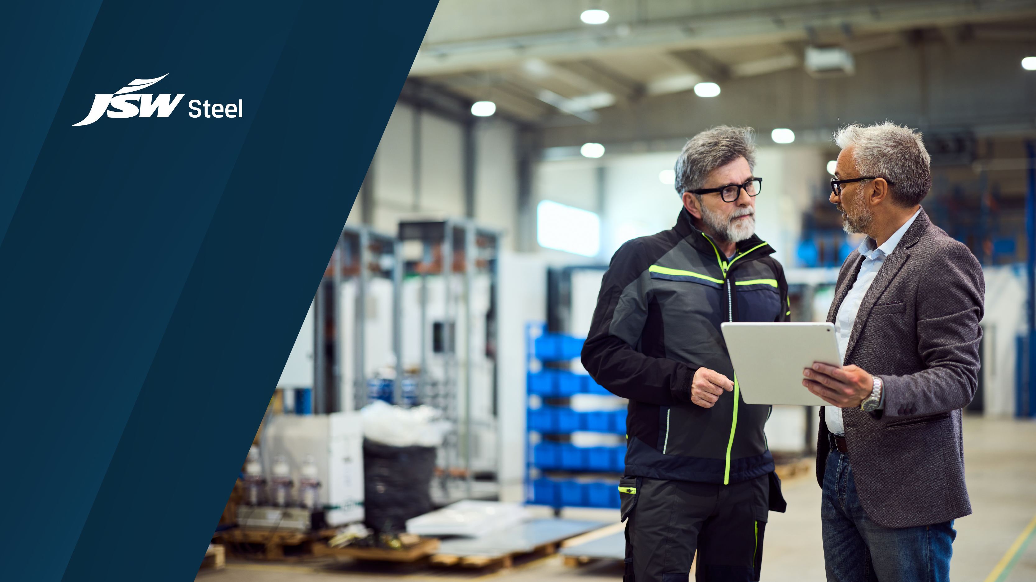 A wide image showing two men in a manufacturing facility discussing information on a tablet, with the JSW Steel logo displayed on a dark blue graphic overlay on the left.