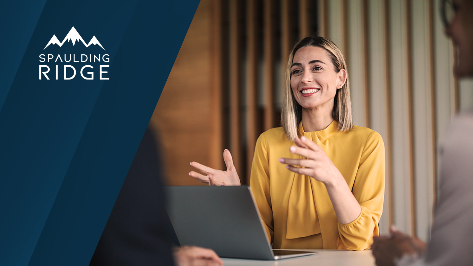 Spaulding Ridge logo displayed. A professional woman in a yellow top speaks and gestures while seated at a table with a laptop during a business meeting.