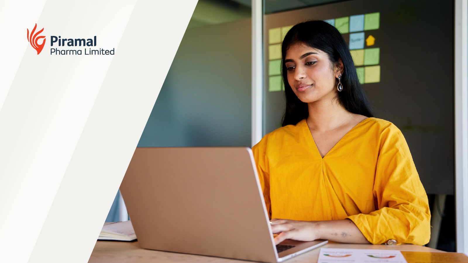 A woman working on a laptop at a desk in a modern office, with the Piramal Pharma Limited logo displayed on the left side of the image.