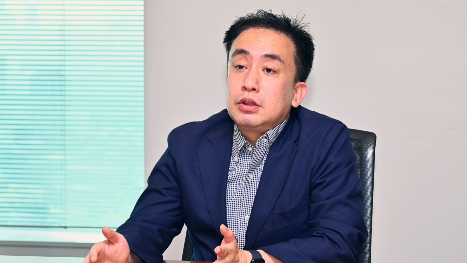 Godiva Japan Customer Story Illustrative Image 1 Ein Mann in einem Blazer und einem karierten Hemd sitzt an einem Schreibtisch und unterstreicht seine Rede mit Gesten. Ein Fenster mit geschlossenen Läden befindet sich im Hintergrund.