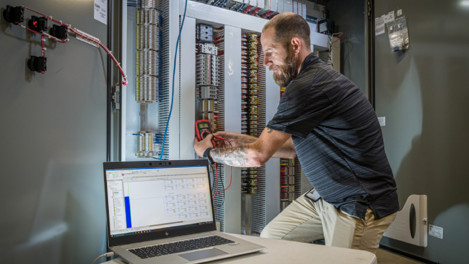 Technician using a multimeter to test a large control panel, with a laptop displaying system diagnostics.