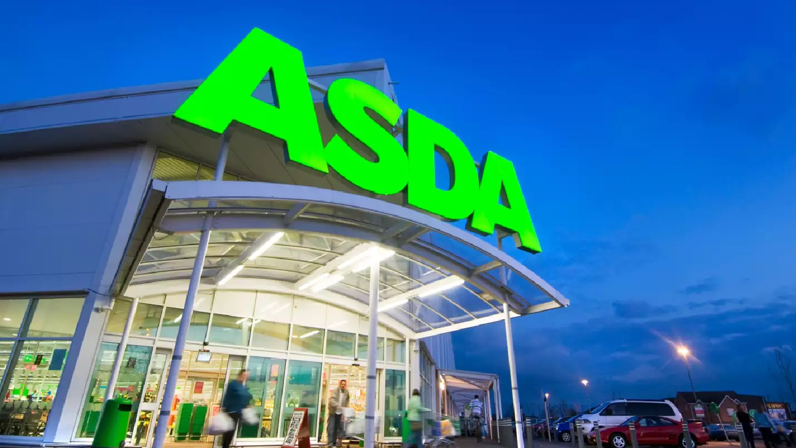The exterior of an Asda supermarket at dusk, with the store’s bright green logo illuminated above the glass entrance as shoppers enter and exit.