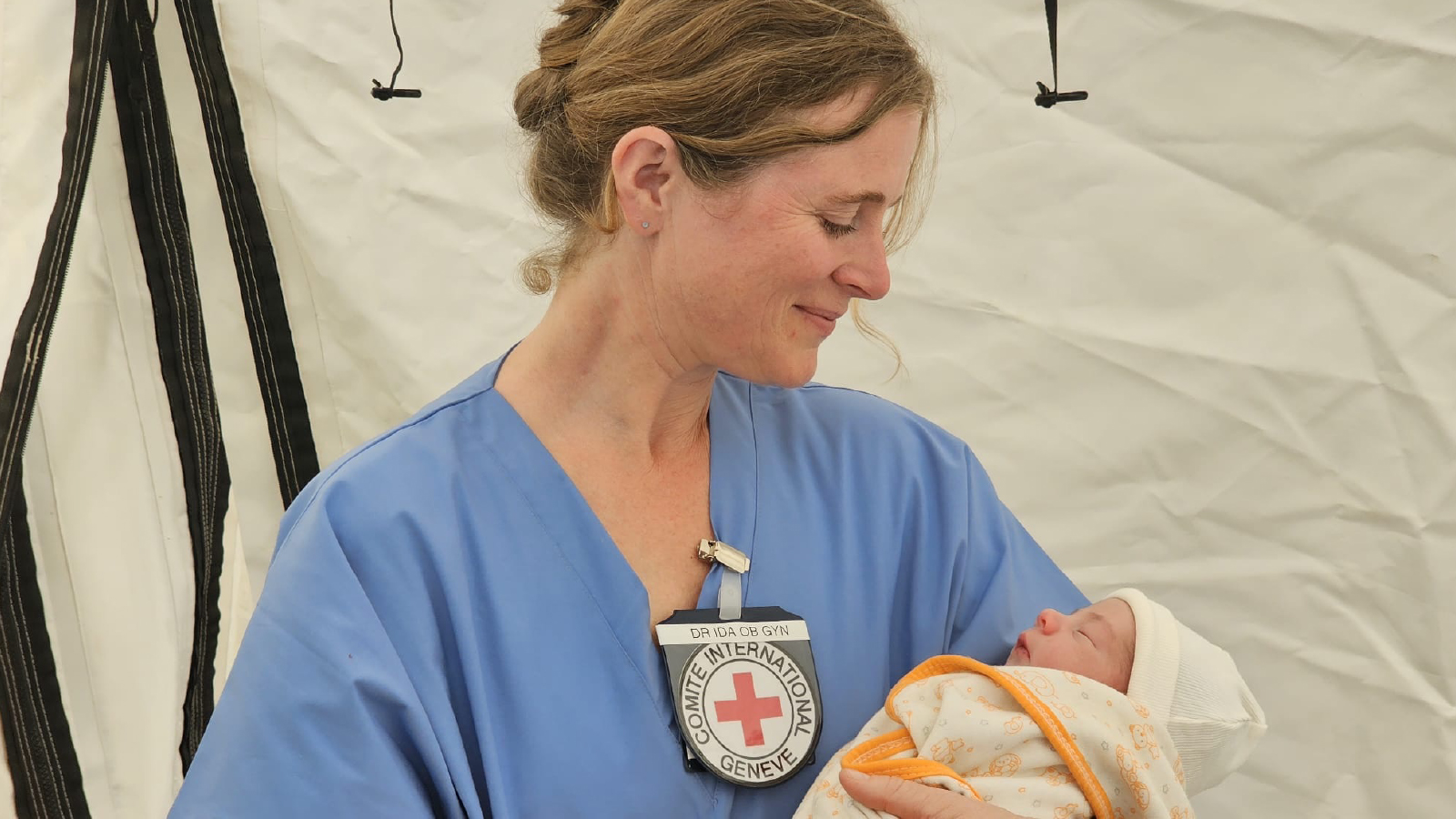 An ICRC medical worker in blue scrubs smiles while holding a newborn baby wrapped in a blanket inside a tent.